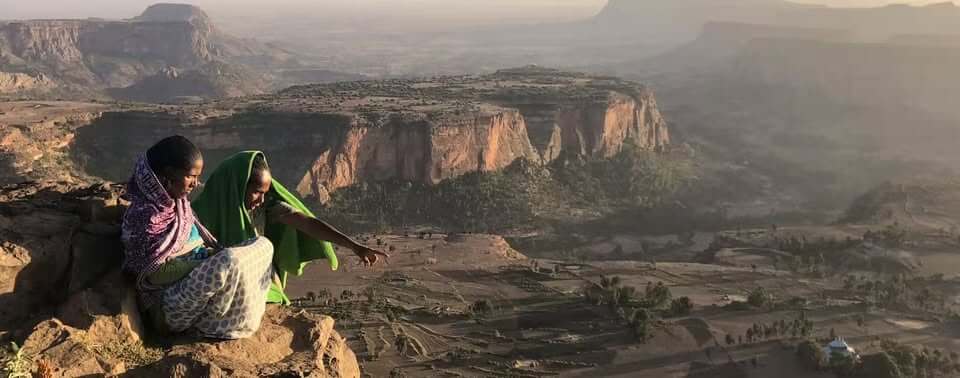 Two Ethiopian women  sitting at the edge of a mountain looking down on the country, one of them pointing at something interesting.